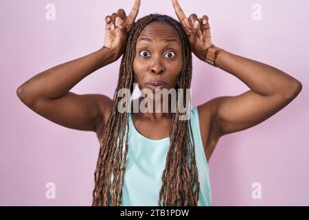 afroamerikanische Frau, die über rosa Hintergrund steht und lustige Gesten mit dem Finger über dem Kopf wie Stierhörner macht Stockfoto