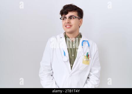 Junger, nicht binärer Mann, der Ärzteuniform und Stethoskop trägt und mit einem Lächeln im Gesicht zur Seite blickt, natürlicher Ausdruck. Lacht selbstbewusst. Stockfoto