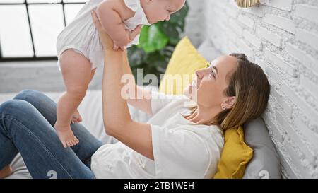 Fröhliche Mutter, die auf dem Bett liegt, selbstbewusst die glückliche Tochter in der Luft hält, Spaß hat, während sie lächelt und zusammen in einem warmen, gemütlichen Schlafzimmer spielt. Stockfoto