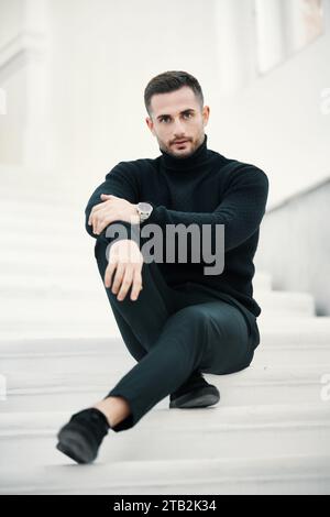 Das modische Herrenmodell sitzt selbstbewusst auf einer weißen Treppe, in schwarzer Kleidung Stockfoto