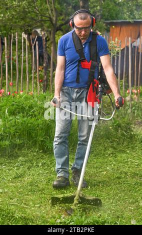 Ein Landwirt mäht das hohe grüne Gras mit einem Rasentrimmer. Stockfoto