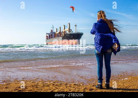 Eine Frau filmt auf ihrem Handy ein großes Frachtschiff an der Küste, das auf Grund gelaufen ist, und einen Kitesurfer. Vityazevo, Russland - 12.02.2023 Stockfoto