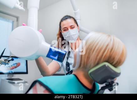 Zahnärztlicher Klinikpatient besucht moderne medizinische Station. Zahnarztärztin gekleidete Uniform-Zeigezahnärztliche Röntgenmaschine. Patient sitzt im Stuhl. Gesundheit Stockfoto