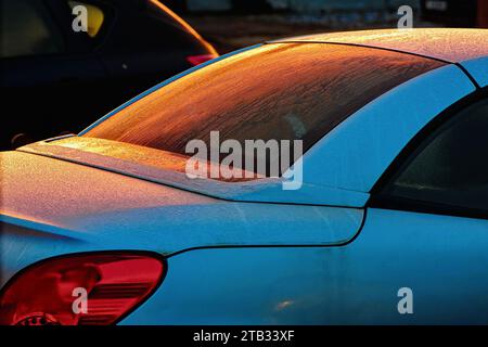 Nahaufnahme eines silbernen Autos mit Reflexen des roten Sonnenlichts auf der Heckscheibe, die von Frost und Regentropfen bedeckt ist und eine abstrakte Form schafft Stockfoto