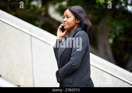 Junge Schwarze Karrierefrau, die draußen eine Treppe raufgeht, während sie telefoniert Stockfoto