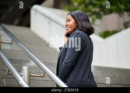 Junge Schwarze Karrierefrau, die draußen eine Treppe raufgeht, während sie telefoniert Stockfoto