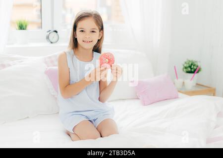 Glückliches junges Mädchen, das einen Donut in einem hellen und gemütlichen Schlafzimmer hält Stockfoto