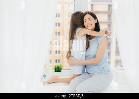 Mutter und Tochter teilen sich eine warme Umarmung am Fenster und strahlen Liebe und Glück aus Stockfoto