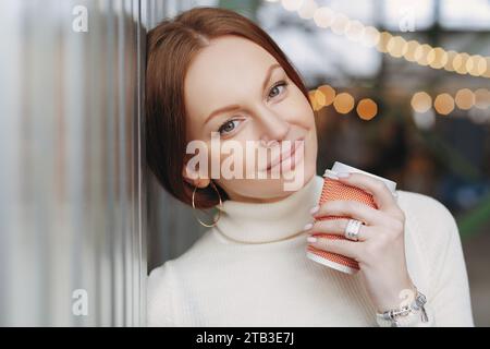 Rothaarige Frau mit Kaffee, die sanft am Fenster lächelt Stockfoto