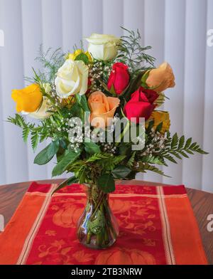 Wunderschöner Strauß aus farbigen Rosen - rot, weiß, pfirsichfarben, gelb, in einer Glasvase auf einem hübschen orangen Tischläufer. Stockfoto