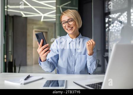 Eine junge Geschäftsfrau, freiberuflich im Büro mit einem Laptop am Schreibtisch sitzend, auf das Handy blickend, mit einer Siegesgesgestik, glücklich und lächelnd. Stockfoto
