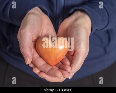 Eine ältere Frau hält eine rote Herzform in ihren Handflächen. Nahaufnahme. Stockfoto