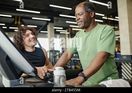 Ein älterer schwarzer Mann, der im Fitnessstudio auf dem Laufband läuft, mit einem jungen kaukasischen Athleten neben ihm Stockfoto