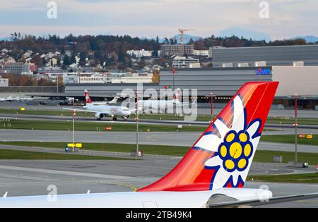 Vertikaler Stabilisator eines Airbus der Schweizer Ferienfluggesellschaft Edelweiss Air, dahinter zwei Flugzeuge der Swiss International Air Lines auf der Landebahn Stockfoto