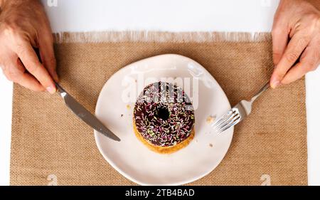 Blick aus einem hohen Winkel auf den Schokoladen-Donut mit Streuseln auf dem Teller. Donut auf Platte und Gabel und Messer in Händen. Stockfoto