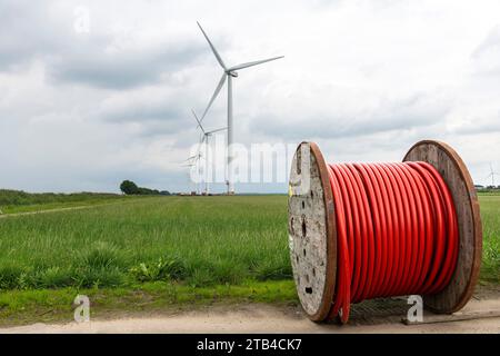 Niedriger Winkel auf ein großes Rad oder eine Spule mit rotem Kabel vor einer langen Reihe moderner Windturbinen oder Windmühlen auf typisch niederländischem Landwirtschaftsland Stockfoto