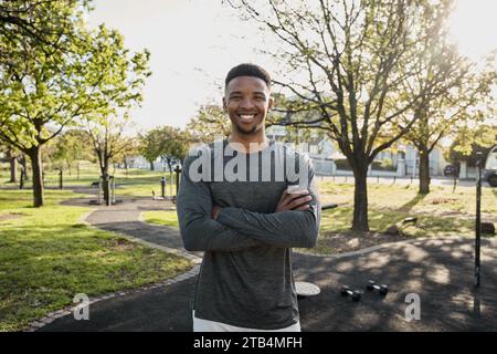 Glücklicher junger schwarzer Mann in Sportbekleidung, der mit überkreuzten Armen im Park in die Kamera schaut Stockfoto