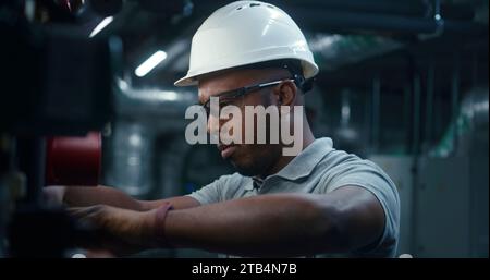 Nahaufnahme eines afroamerikanischen Schwerindustriearbeiters in Sicherheitsuniform und Schutzhelm, der das Energiesystem inspiziert und justiert. Professioneller Ingenieur arbeitet an modernen Fertigungs- oder Industrieanlagen. Stockfoto