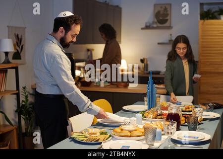 Seitenansicht eines bärtigen jüdischen Mannes in einem Totenkopf-Serviertisch zum Abendessen, während er sich auf die Feier von Hanukka mit seiner Familie zuhause vorbereitet Stockfoto