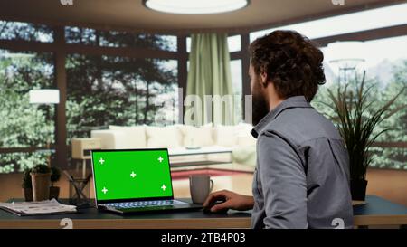 Wohlhabender Investor entspannt sich in seinem eleganten Berghütte, während er sich die Greenscreen-Anzeige auf seinem Notebook anschaut. Der ausländische Supervisor arbeitet im wohlhabenden Leben mit einem isolierten Mockup-Panel auf einem Laptop. Stockfoto