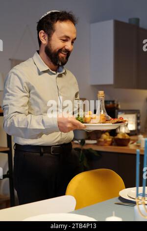 Ein lächelnder bärtiger Mann hält einen Teller mit einer Flasche Öl und hausgemachten Snacks, während er einen Tisch für das Hanukkah-Abendessen in der Küche serviert Stockfoto