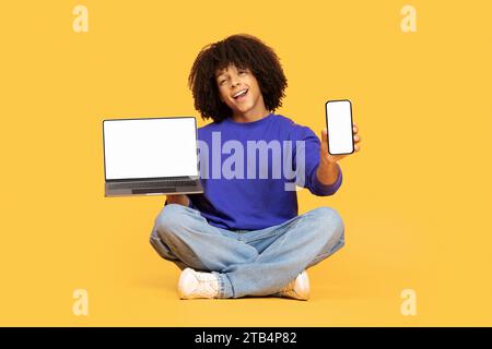 Schöner junger schwarzer Mann, der Laptop und Smartphone mit leeren Bildschirmen zeigt Stockfoto
