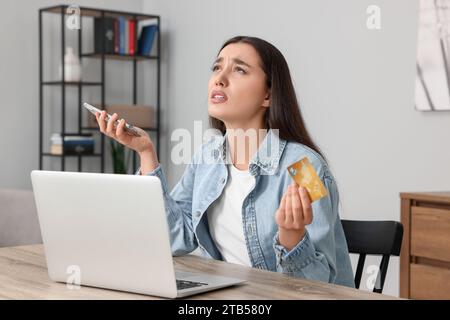 Verwirrte Frau mit Kreditkarte, Smartphone und Laptop am Tisch drinnen. Seien Sie vorsichtig – Betrug Stockfoto