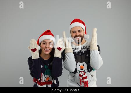 Junges Paar in Weihnachtspullover, Weihnachtsmannmützen und gestrickten Fäustlingen auf grauem Hintergrund Stockfoto