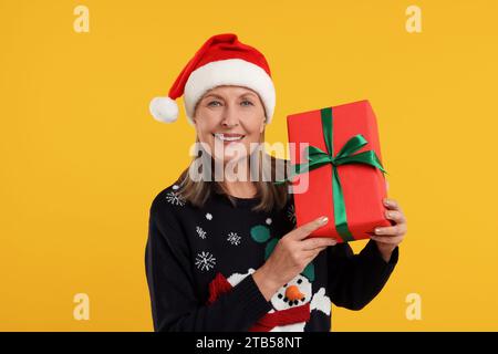 Glückliche ältere Frau im Weihnachtspullover und Weihnachtsmann-Hut mit Geschenk auf orangem Hintergrund Stockfoto