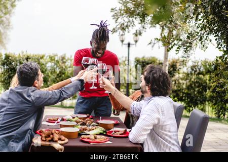 Verschiedene Freunde lachen und toasten mit Rotwein an einem Tisch im Freien, gefüllt mit köstlichem Essen. Stockfoto