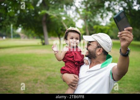 Wunderschöner junger indischer Vater mit Hut und Sonnenbrille, der mit seinem kleinen Mädchen im Park oder Garten tanzt. Stockfoto