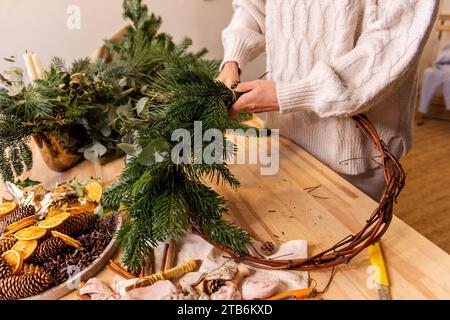 Draufsicht Komposition weiblicher Hände, die Weihnachtskranz aus natürlichen Zweigen von Fichte, Kiefer, Eukalyptus herstellen. Das Mädchen wickelt grüne Zweige auf die Kraft-Basis Stockfoto