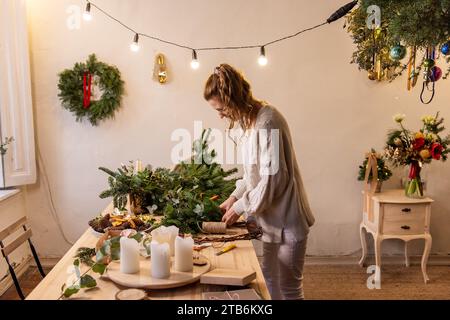 Die junge Frau macht zu Hause einen handgefertigten Weihnachtskranz aus natürlichen Materialien. Authentisches Apartment mit skandinavischem Interieur. DIY-Anweisung von flori Stockfoto