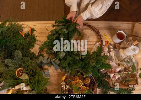 Draufsicht Komposition weiblicher Hände, die Weihnachtskranz aus natürlichen Zweigen von Fichte, Kiefer, Eukalyptus herstellen. Das Mädchen wickelt grüne Zweige auf die Kraft-Basis Stockfoto