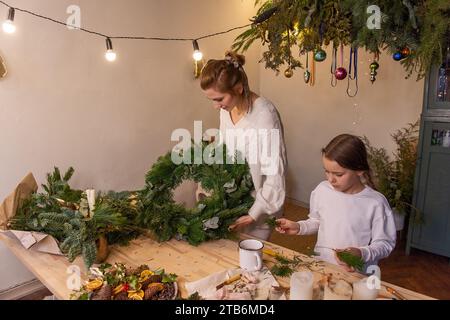 Junge Mutter und kleine Tochter machen zusammen Weihnachtskranz aus Fichte, Eukalyptuszweigen. Alleinerziehende Mutter zeigt Mädchen Tutorial, wie man Neues macht Stockfoto