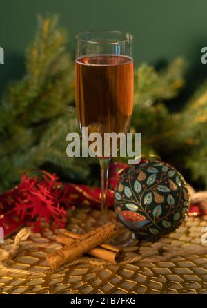 Hohes Glas Champagner vor grünem Hintergrund mit Weihnachtsbaumlaub, Glaskugeln und Zimtstangen mit rotem Band auf goldenem Tisch Stockfoto
