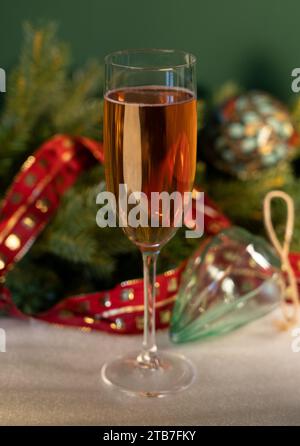 Hohes Glas Champagner vor grünem Hintergrund mit Weihnachtsbaumlaub, Glaskugeln mit rotem Band auf weißem Stoff Stockfoto