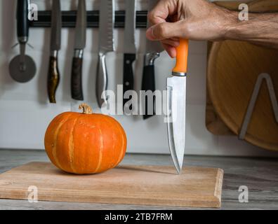 Kürbis auf einem Schneidebrett und ein Messer in der Hand eines Mannes vor einem Hintergrund von Messern an einer weißen Wand Stockfoto