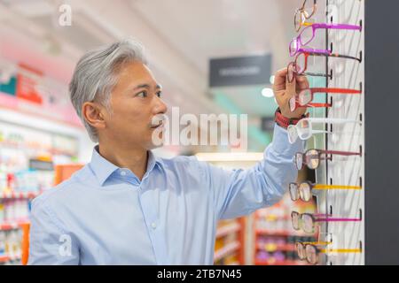 Schöner ruhiger älterer Mann mit grauen Haaren in Hemd, der Brillen auswählt, während er beim Einkaufen in einem optischen Geschäft in der Nähe eines Gepäckträgers steht Stockfoto