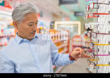 Schöner ruhiger älterer Mann mit grauen Haaren in Hemd, der Brillen auswählt, während er beim Einkaufen in einem optischen Geschäft in der Nähe eines Gepäckträgers steht Stockfoto