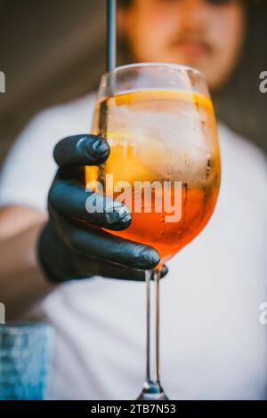 Nicht erkennbarer Barkeeper mit Handschuhen, die einen orangen Cocktail mit Eis halten und einen Strohhalm an der Bar in unscharfem Hintergrund stehen Stockfoto