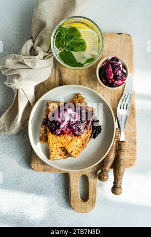 Frisch zubereitete Crepes werden mit einem Klecks Sauerrahm und einem großzügigen Aufstrich Heidelbeermarmelade auf einem rustikalen Holzbrett serviert. Stockfoto