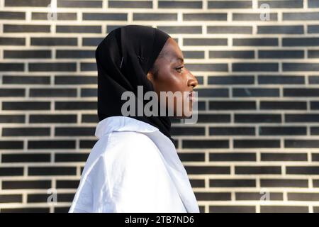 Porträt einer selbstbewussten afroamerikanischen muslimischen Unternehmerin in schwarzem Hijab und weißem formalen, die in der Stadt an einer Ziegelmauer steht Stockfoto