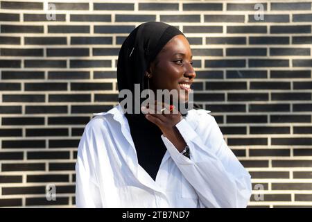 Porträt einer selbstbewusst lächelnden afroamerikanischen muslimischen Unternehmerin in schwarzem Hijab und weißem Formal, die in der Stadt an einer Ziegelmauer steht Stockfoto