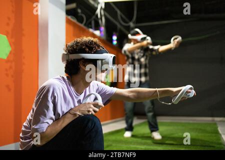 Zwei junge Menschen erleben ein intensives Virtual-Reality-Erlebnis, ausgestattet mit VR-Headsets und Controllern in einer Spielhalle. Stockfoto