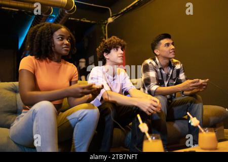 Fokussierte junge multirassische Freunde sitzen auf einer Couch, um an einer lustigen Videospielsession teilzunehmen Stockfoto