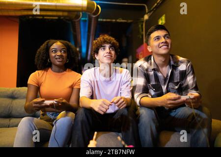 Drei junge multirassische Freunde sitzen auf einer Couch, um ein lustiges Videospiel zu erleben Stockfoto