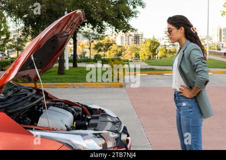 Seitenansicht einer unglücklichen, frustrierten jungen Geschäftsfrau, die mit Händen auf der Hüfte steht und den Motor eines Pannenfahrzeugs am Straßenrand anstarrt Stockfoto