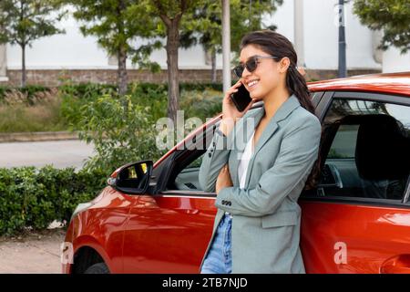 Stilvolle junge, lächelnde Geschäftsfrau in Blazer und Sonnenbrille, die sich über ein Mobiltelefon an ein oranges Auto lehnt Stockfoto