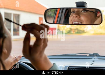 Lächelnde, glamouröse junge kaukasische Geschäftsfrau, die tagsüber in den Rückspiegel blickt und Mascara aufträgt, während sie im modernen Auto sitzt Stockfoto